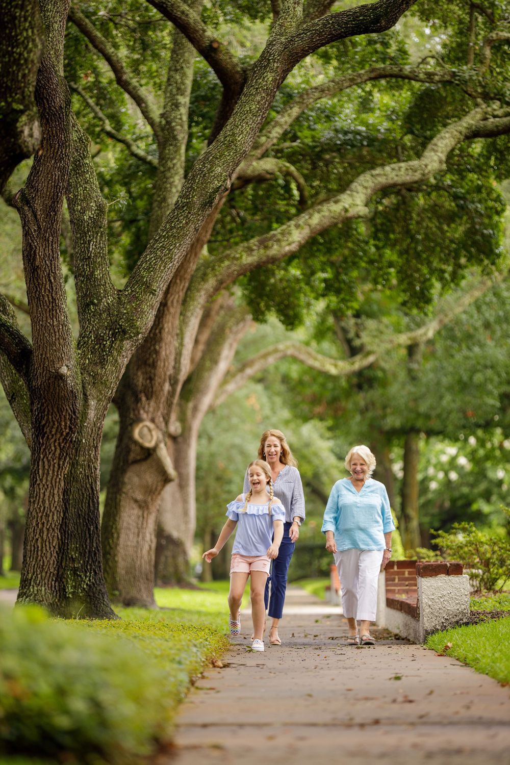Three generations of women walk together along a tree-lined path in a lush green park on a summer day.