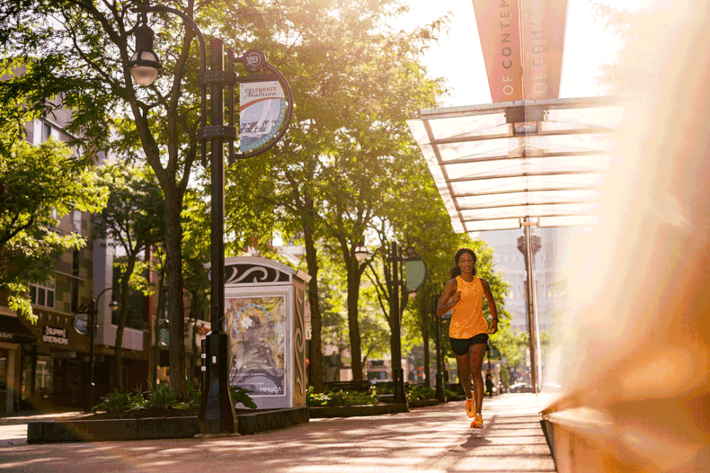 A jogger runs along a sunny tree-lined urban pedestrian street with green trees and modern architecture.