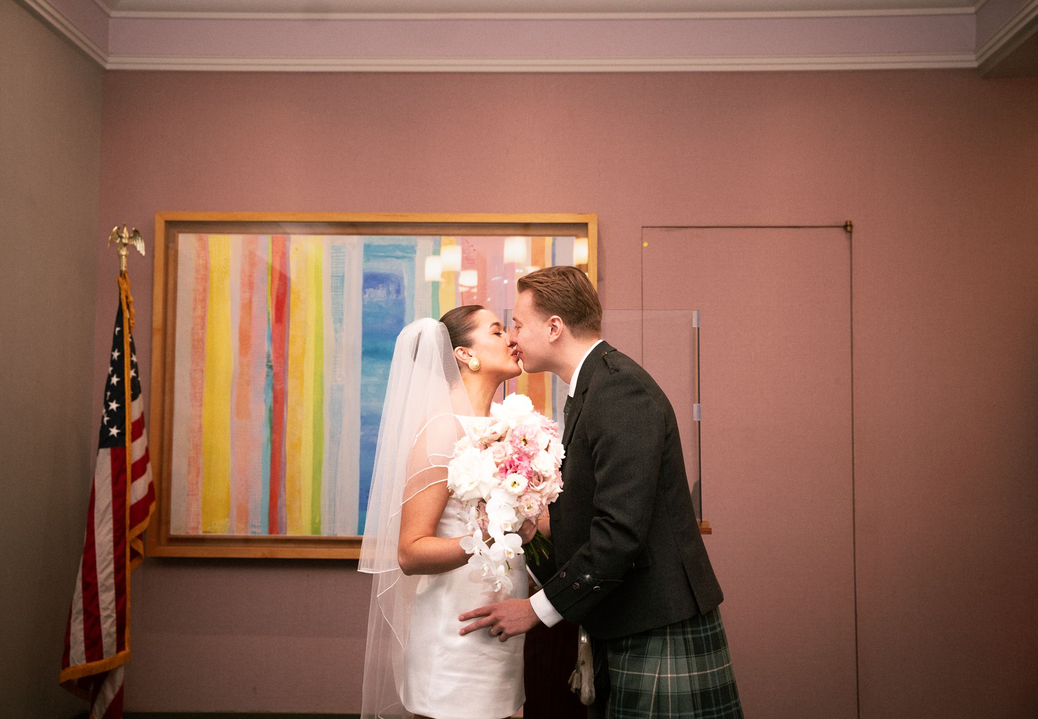 Bride and groom share a kiss at their wedding, with a colorful abstract painting and American flag in the background.