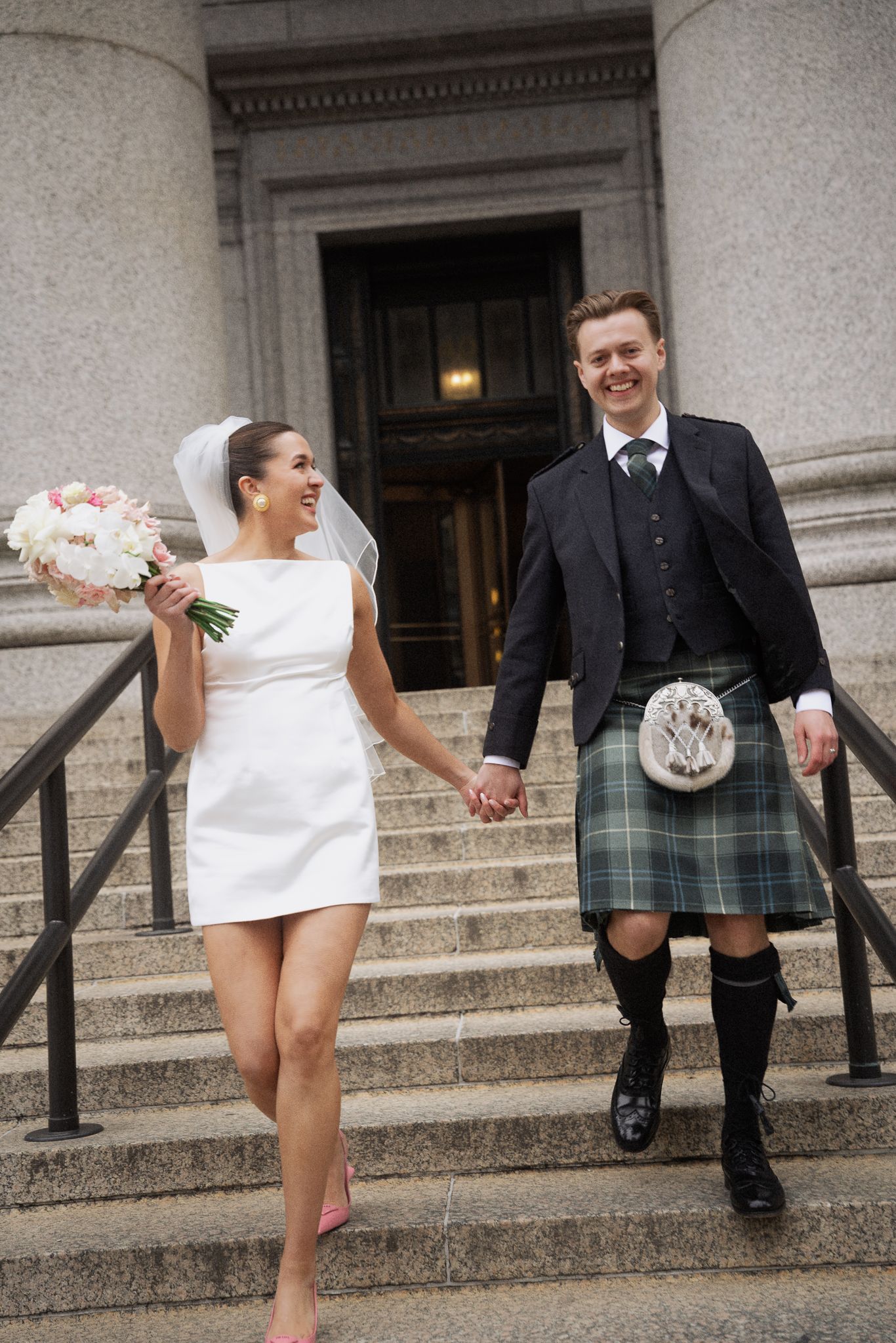 Bride in short white dress and groom in Scottish kilt walk down stone steps holding hands after wedding ceremony.