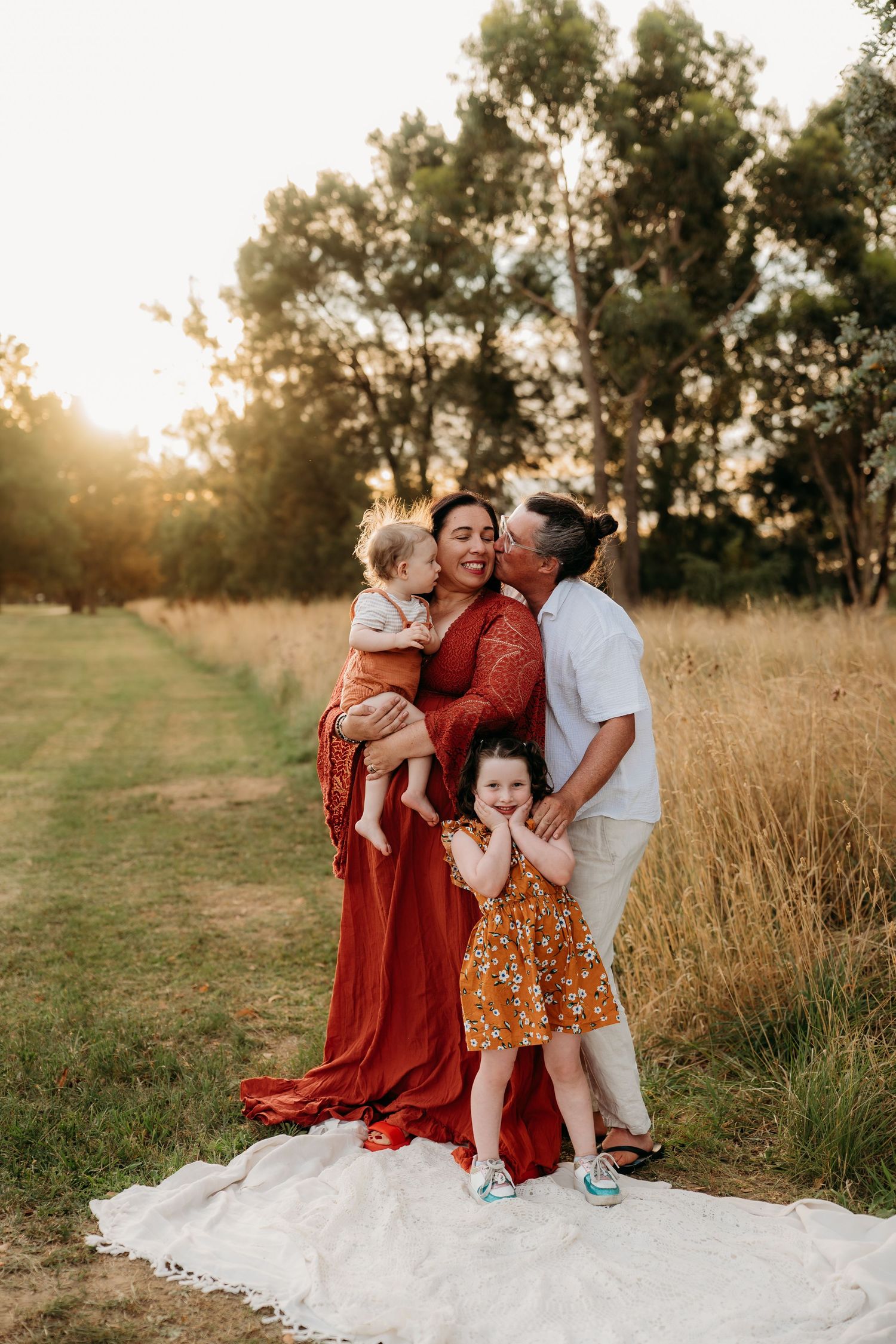 Family of four posing at golden hour in a field, mother in red dress holding baby, father kissing her cheek.