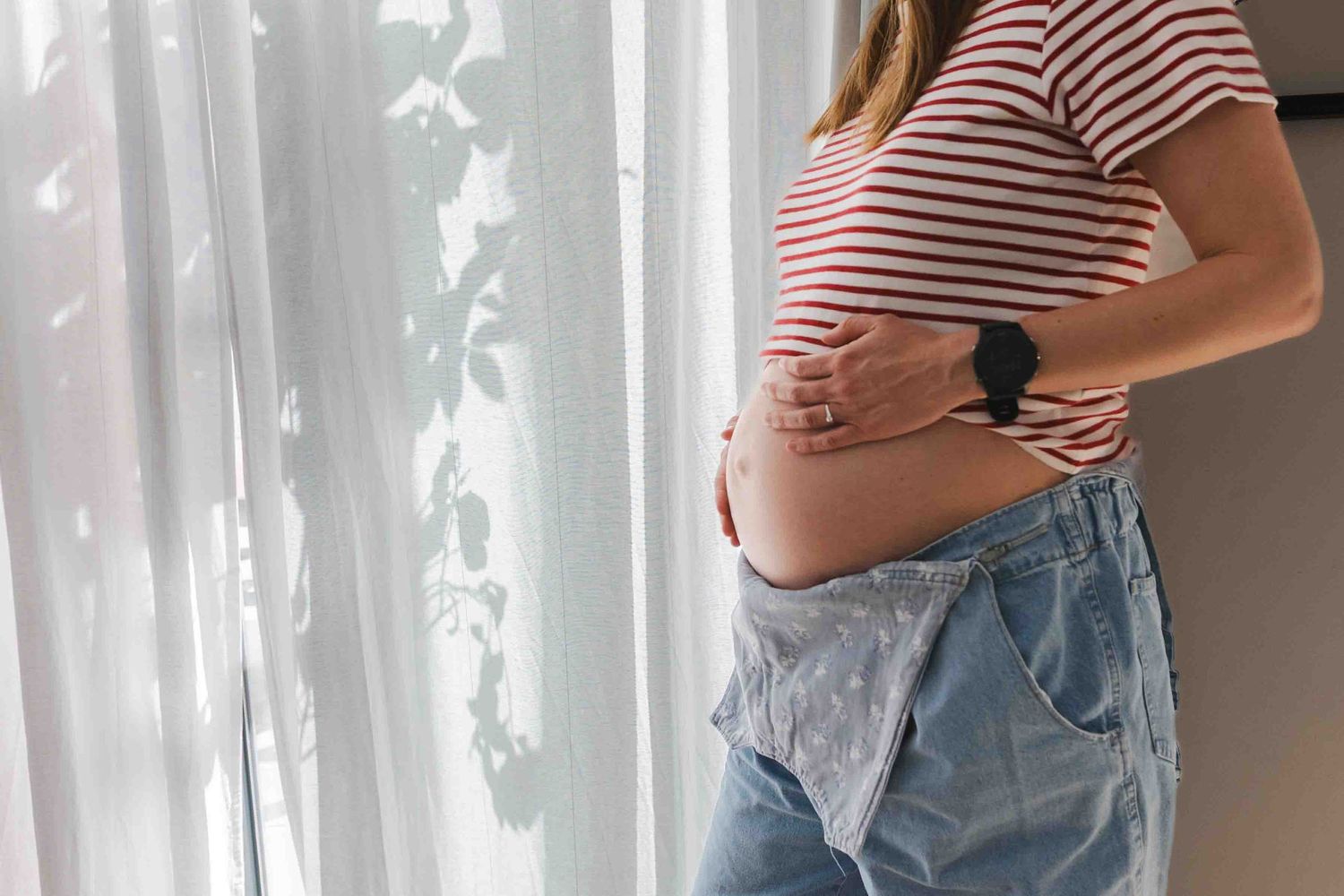 Pregnant woman in striped crop top and jeans holding baby bump near sheer white curtains.