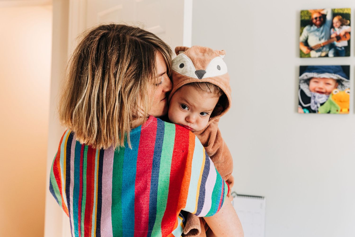 Mother holding baby wrapped in a colorful striped towel with a fox hood, sharing a tender moment at home.
