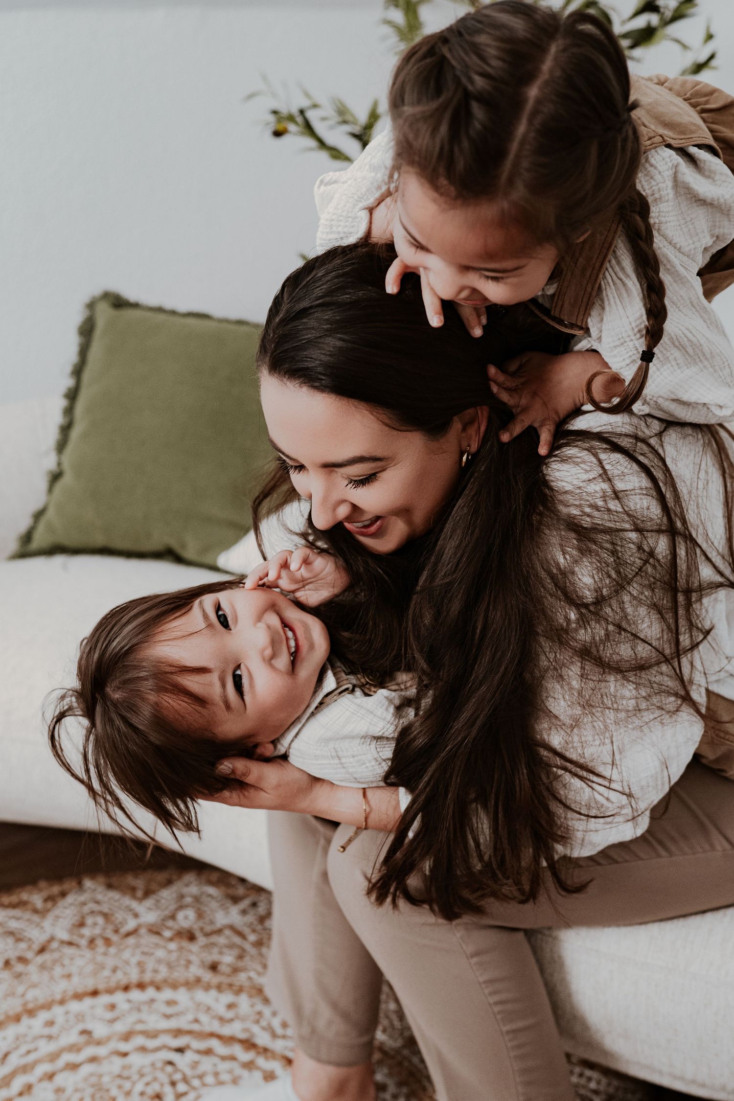 A mother joyfully embraces her two young daughters on a couch in a bright, cozy living room.