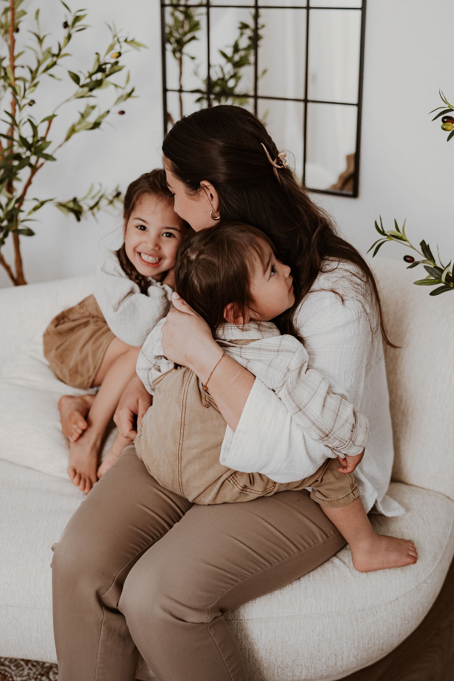 A mother hugs her two young children on a white couch in a bright, plant-filled living room.