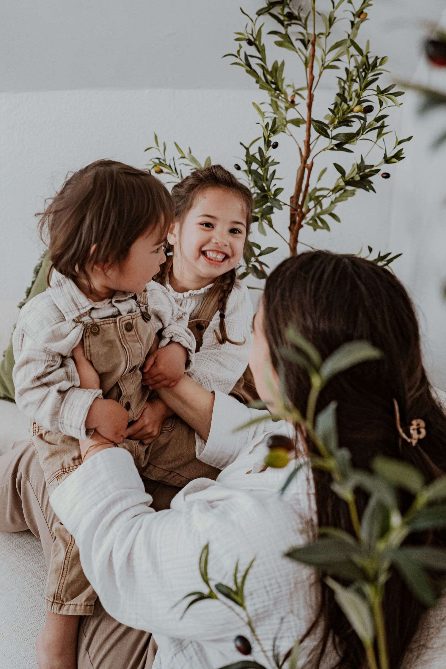 A joyful mother plays with her two young children among green olive branches in a bright, airy room.