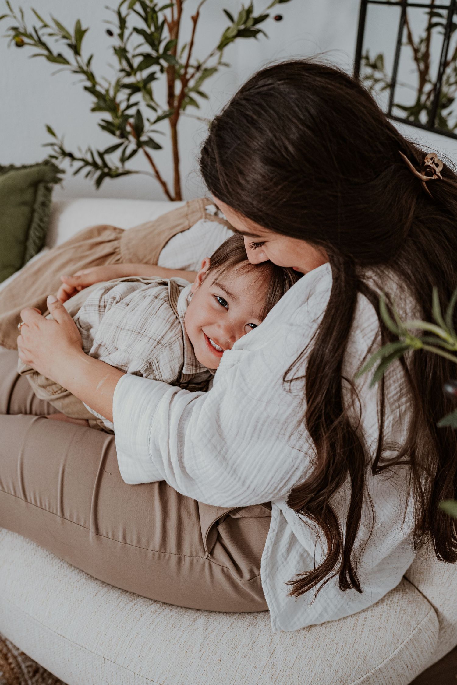 A mother with long dark hair hugs her smiling toddler on a cozy white sofa surrounded by green plants.