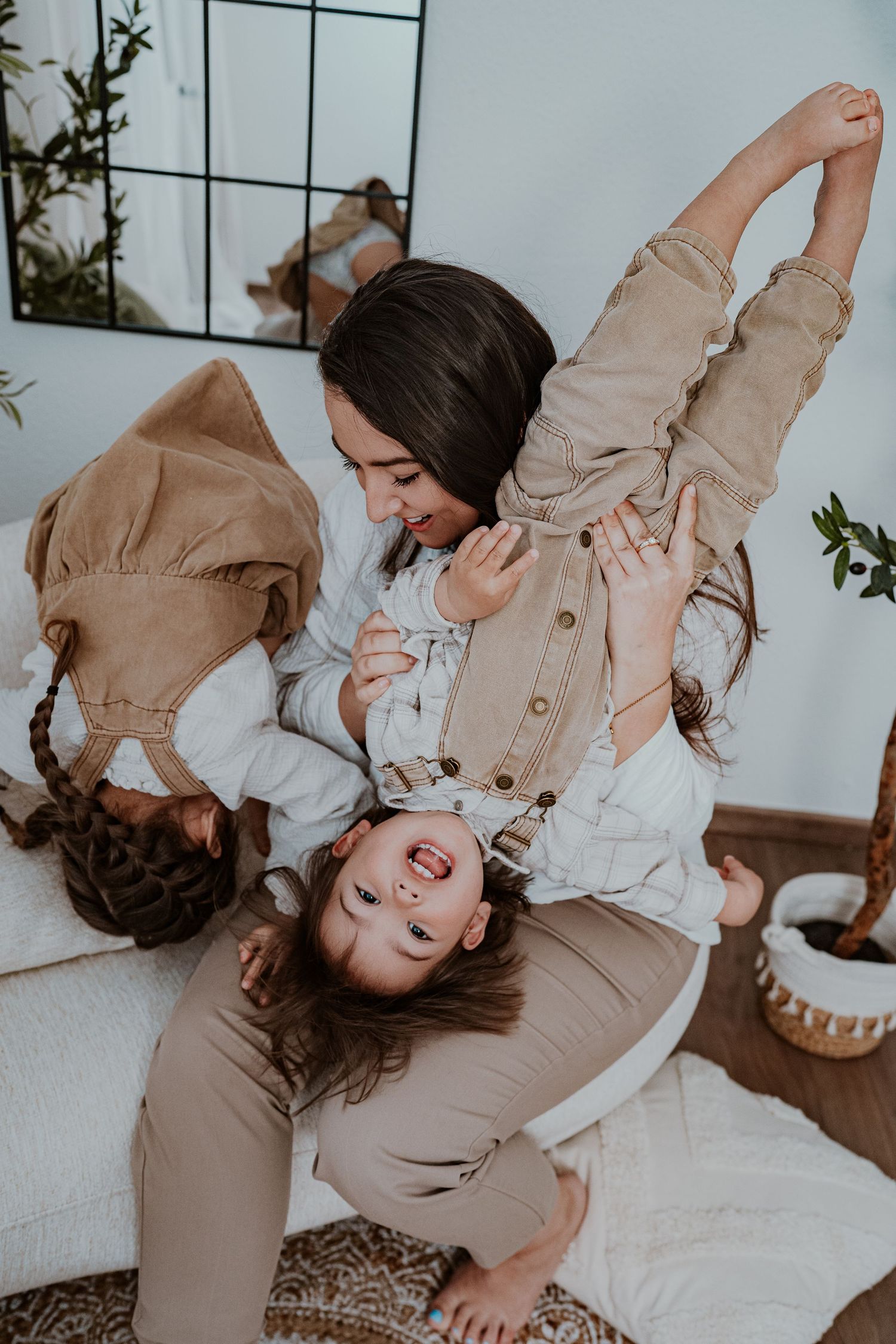 A mother playfully holds her laughing daughter upside down while another child joins the fun on a cozy bed.