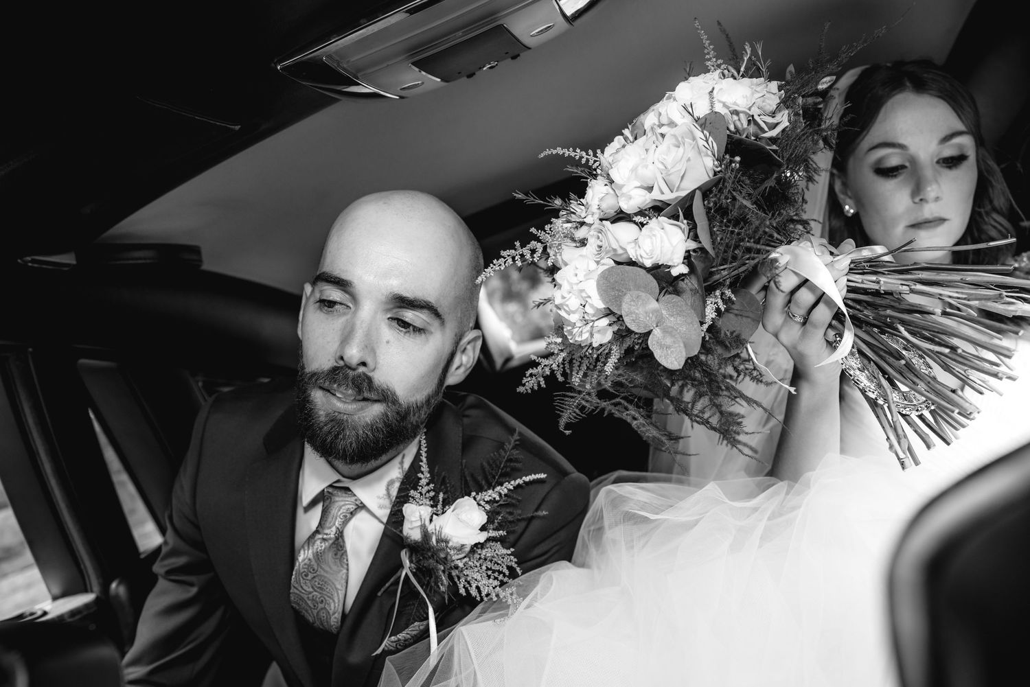 Black and white photo of a bald groom and bride holding a floral bouquet seated together in a car.