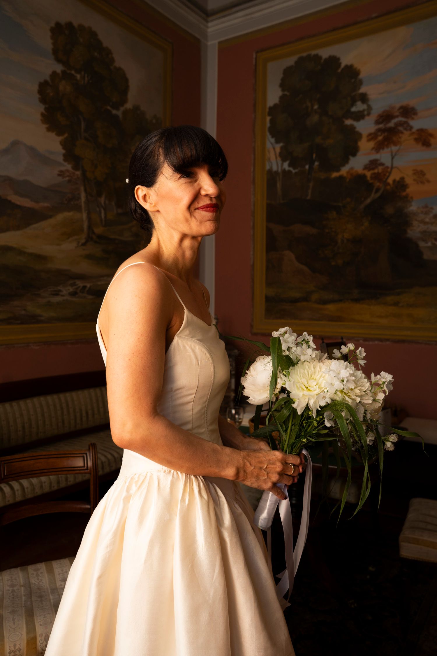Mature bride in ivory gown holding white peony bouquet in room with landscape paintings.