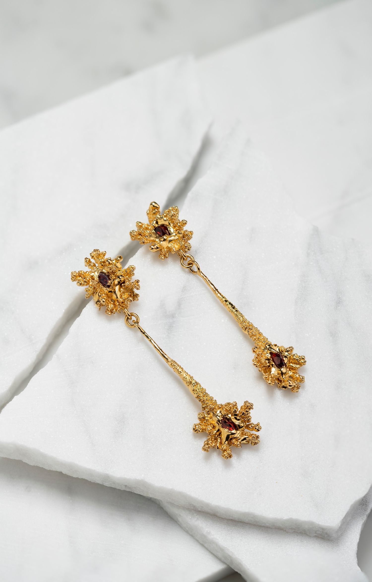 Gold floral drop earrings with red gemstone accents laid on a white marble surface.