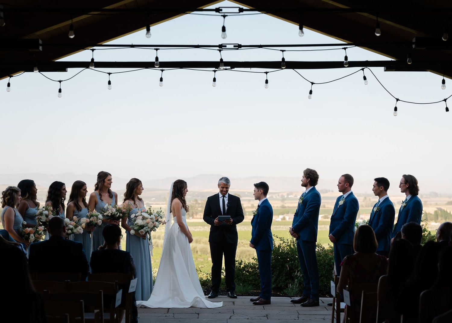 A bride and groom share a romantic moment holding hands in a lush vineyard with rolling golden hills in the background.
