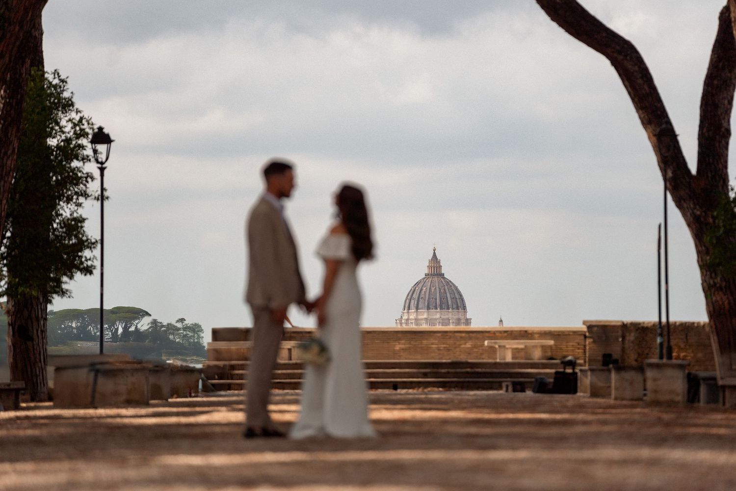 Couple holds hands at Rome's Orangerie Garden with St. Peter's Basilica dome visible in the background.