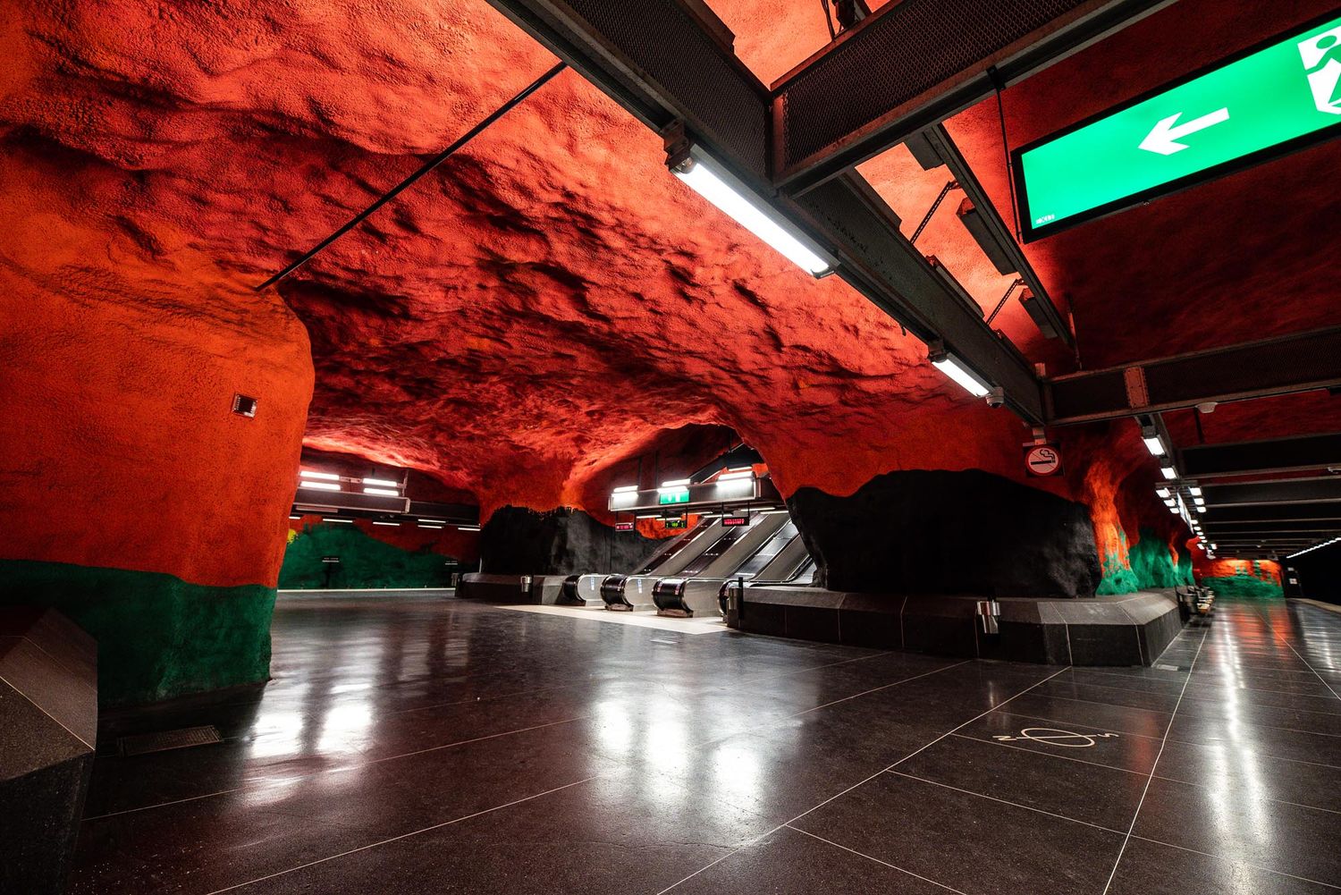 A dramatic red-lit subway station tunnel with modern architectural design features illuminated walkways and exit signs.