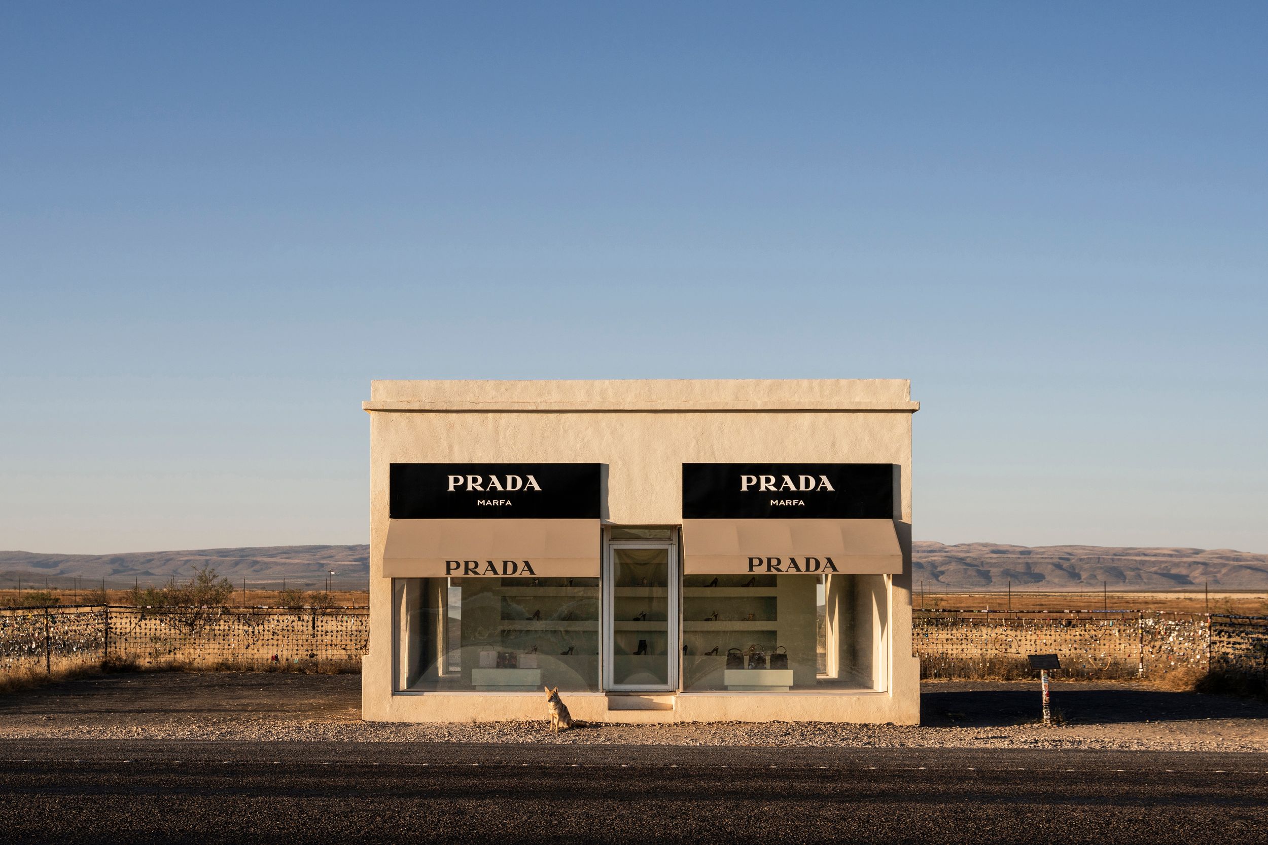 A lone Prada storefront installation stands in the middle of a desert landscape against a clear blue sky.