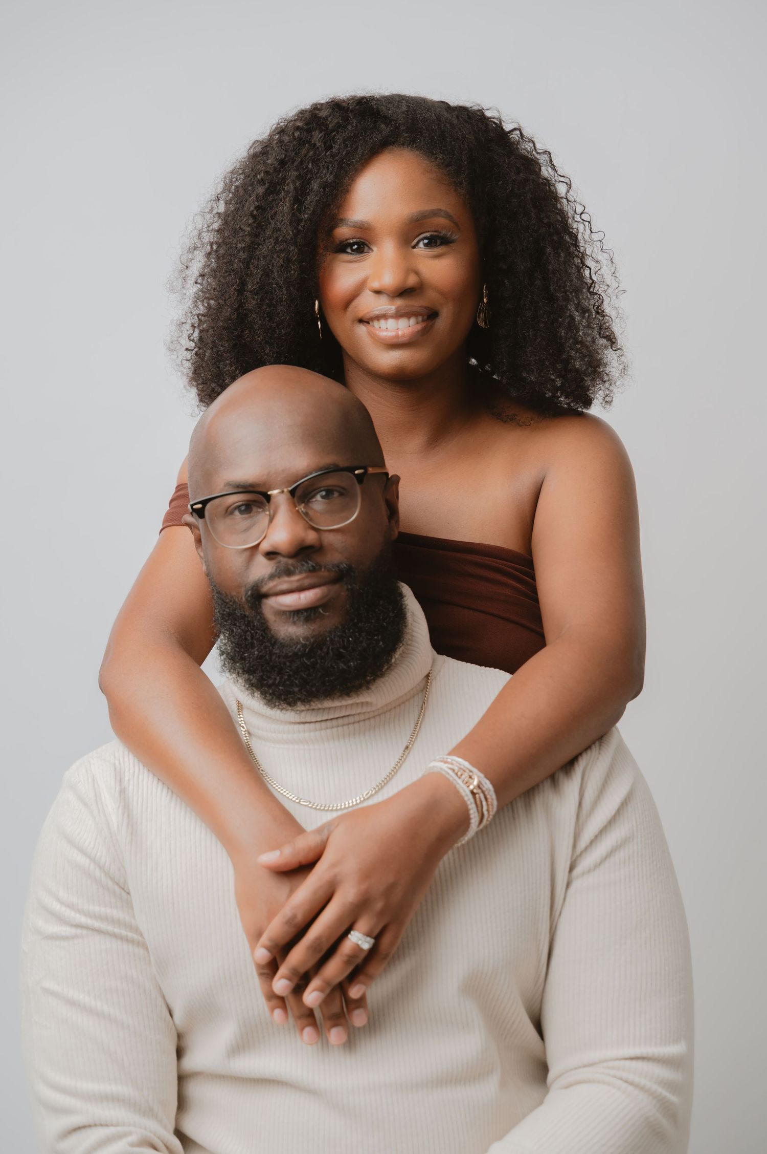 A loving couple in matching cream sweaters embrace against a light gray studio backdrop in a series of romantic poses.