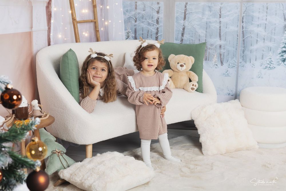 Two small children pose in a cozy white room decorated for Christmas with a teddy bear and throw pillows.