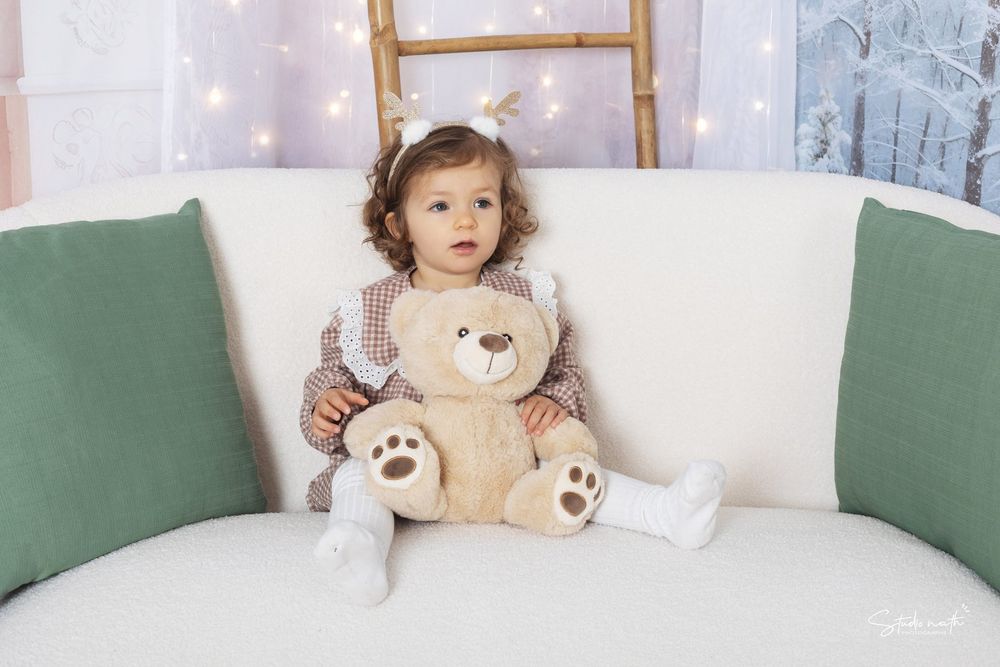 A small child sits between green accent pillows on a white couch holding a plush teddy bear with twinkle lights in background.