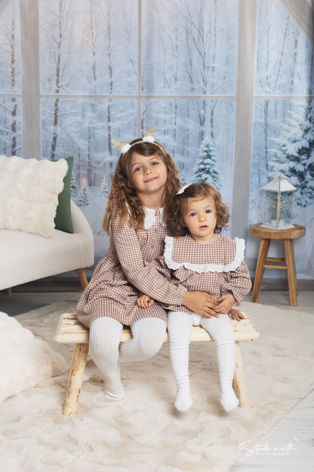 Two children in matching gingham dresses sit together on a bench against a wintry window backdrop.
