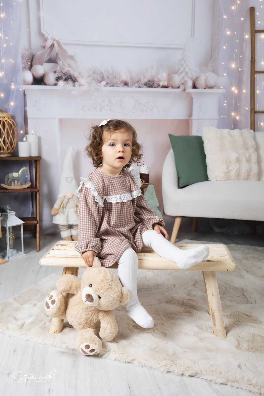 A small child in a brown dress sits on a wooden bench with teddy bears in a cozy holiday-decorated room.