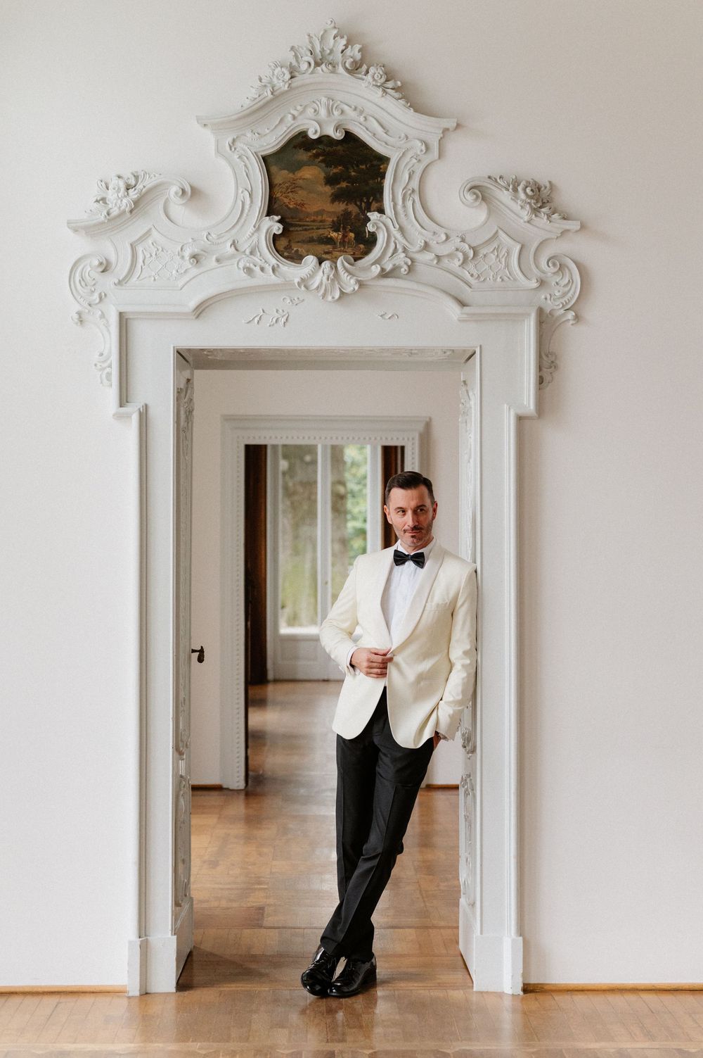 A formal white doorway with ornate baroque molding frames a figure in a white dinner jacket and bow tie.