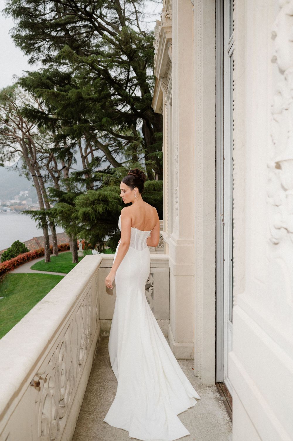 A white wedding dress silhouetted against a balcony overlooking a coastal landscape with trees.