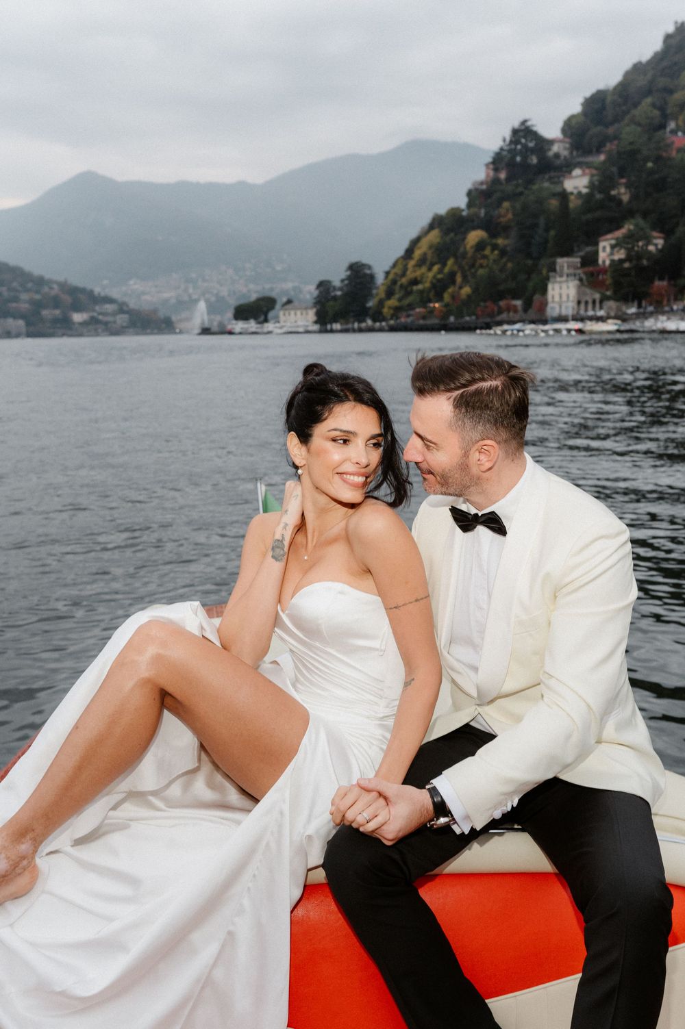 A couple in wedding attire shares intimate moments on a red boat seat with a scenic lake and mountains behind them.