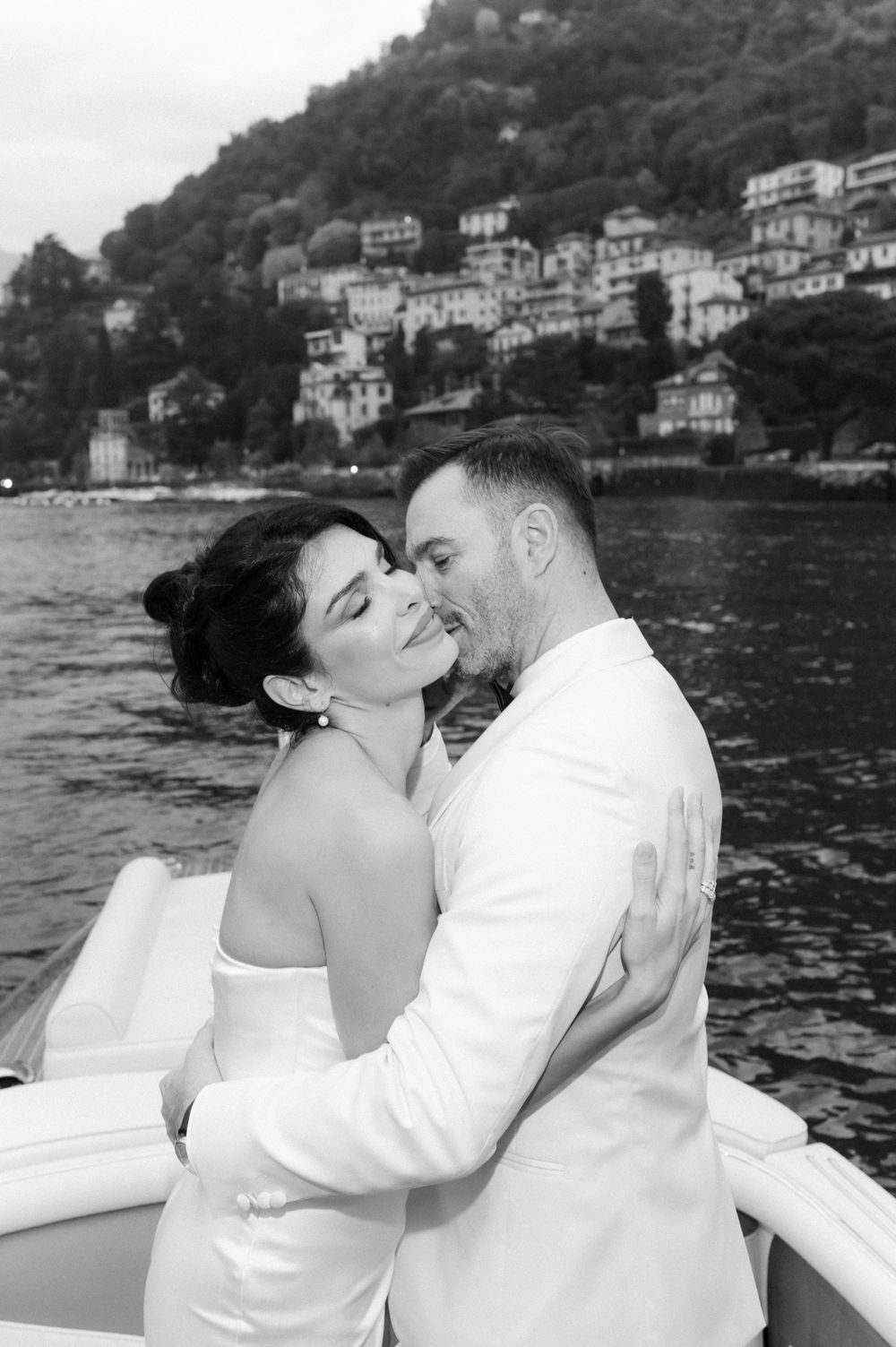 Romantic black and white photos of a couple embracing on a boat with lakeside villas in the background.