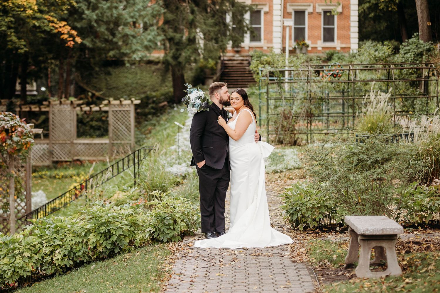 A couple shares an intimate moment in a lush garden setting surrounded by greenery and brick buildings.
