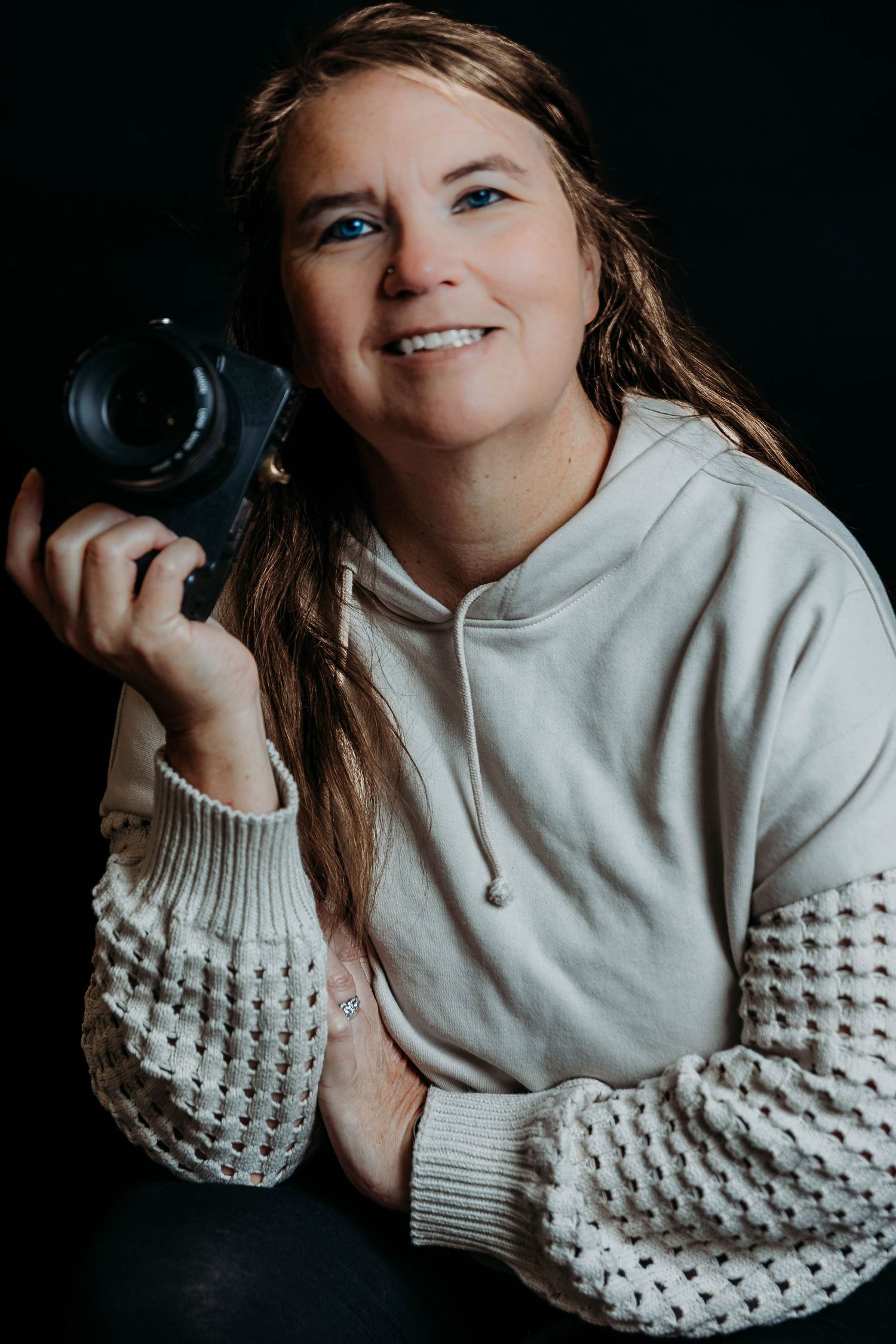 A photographer wearing a cozy white knit sweater poses with a camera against a dark background.