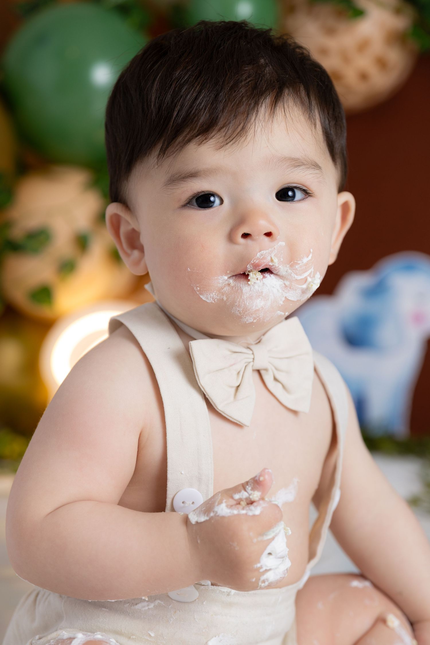 A baby wearing a cream bow tie smiles while covered in cake frosting during a first birthday cake smash photo session.