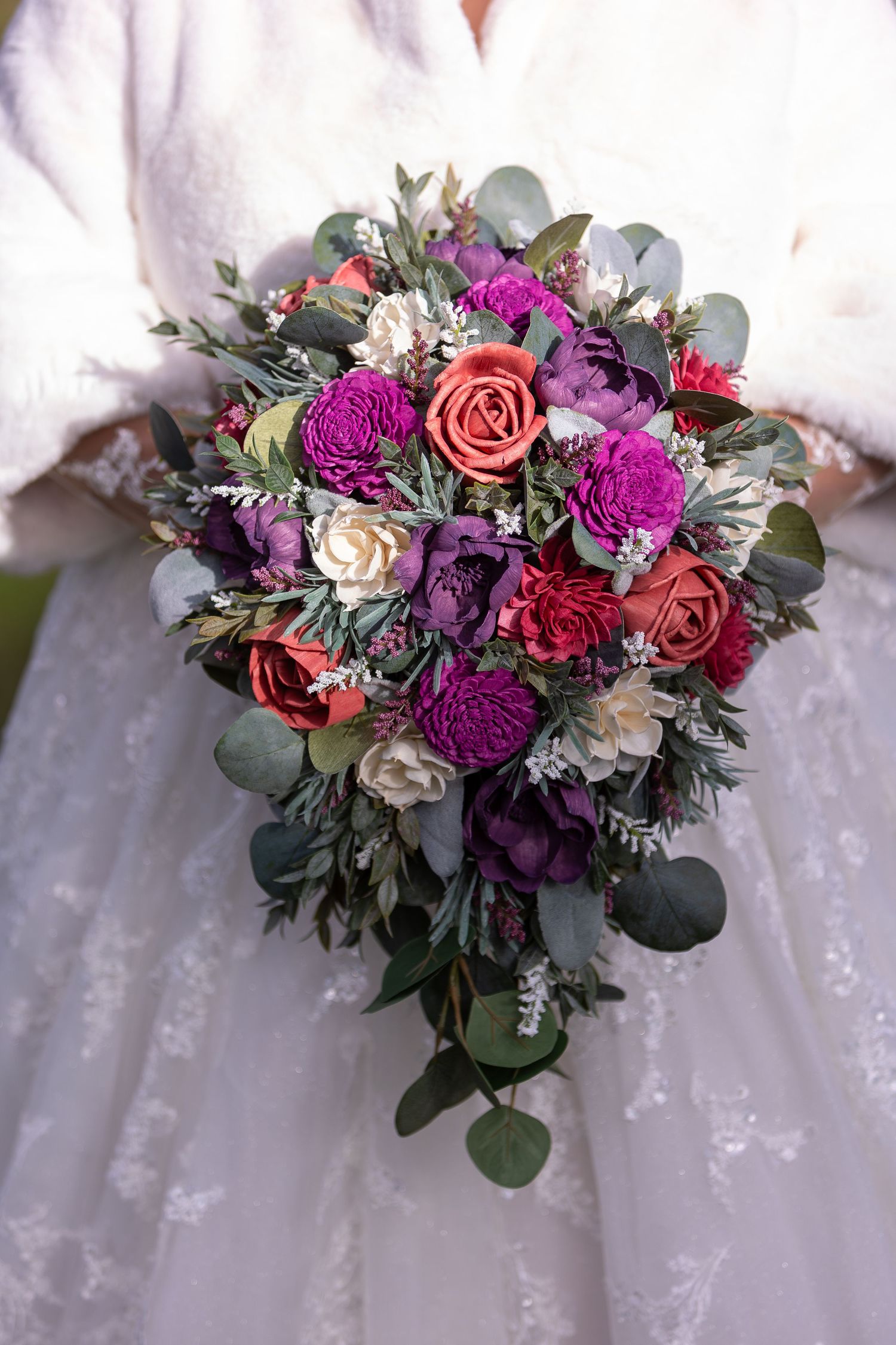 Vibrant bridal bouquet featuring purple carnations, coral roses, and eucalyptus greenery against white lace fabric.