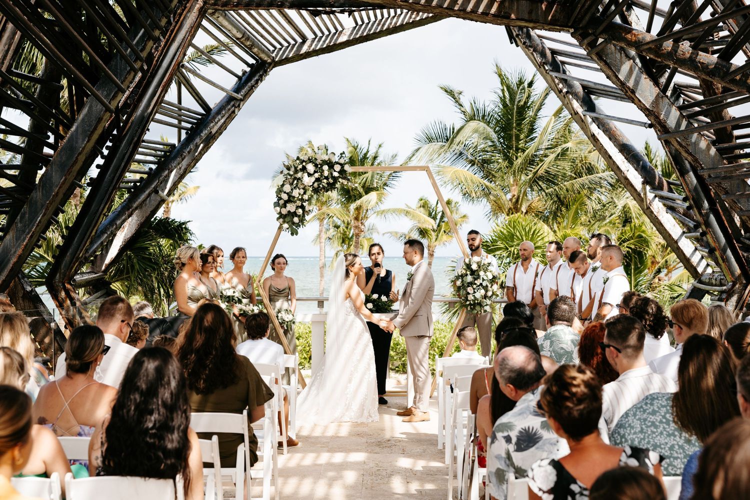 Beach wedding ceremony under a palm frond arch with guests seated on white chairs.