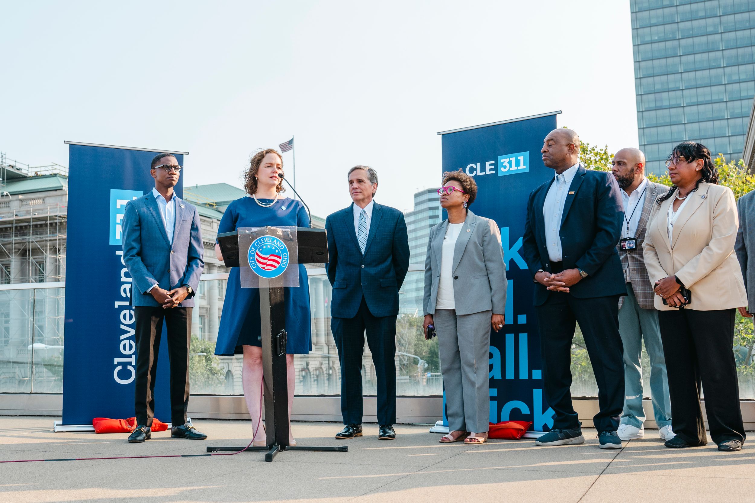 Business professionals gather at a corporate event with blue banners and signage visible in the background.