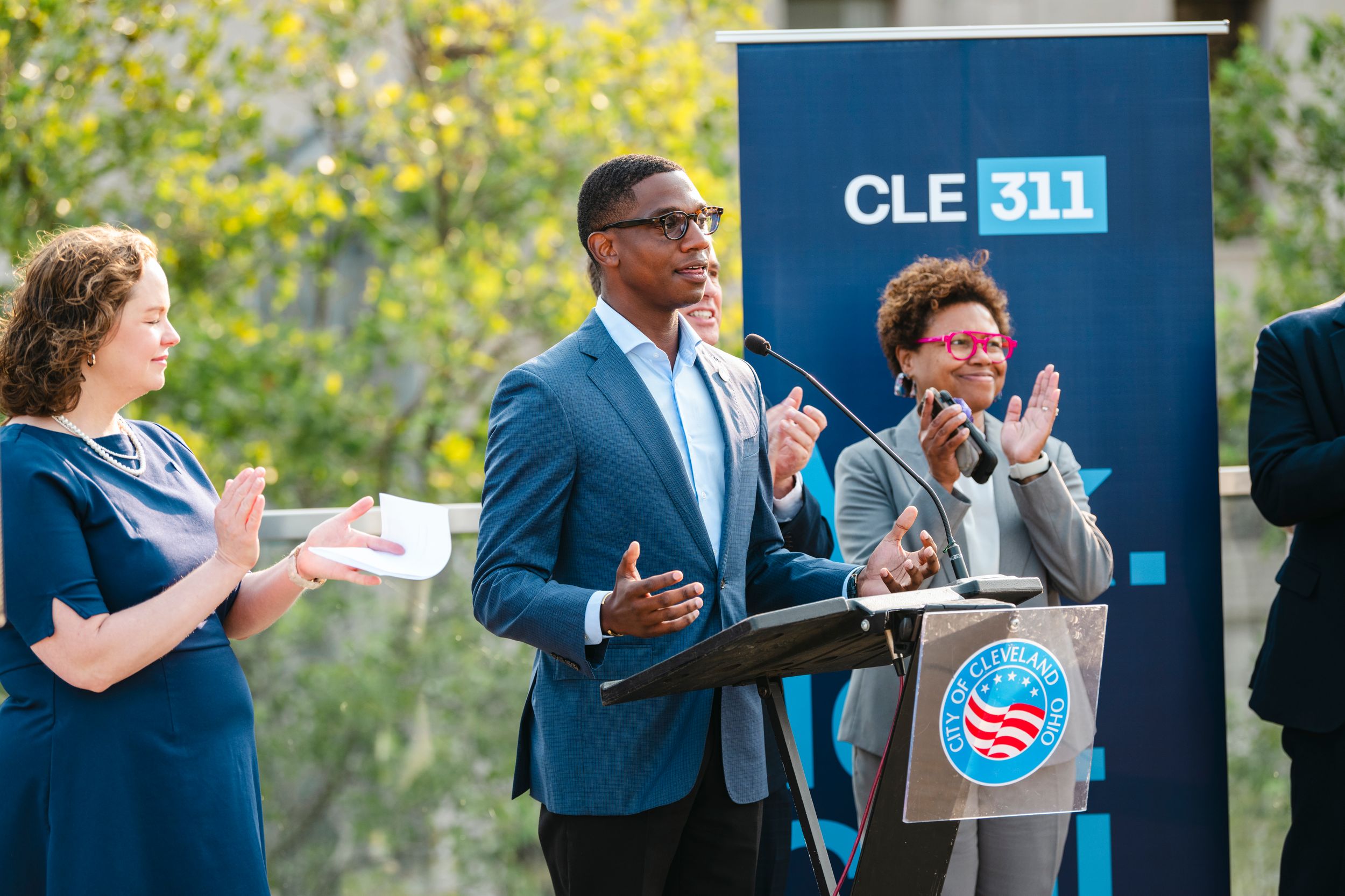 Public speakers at outdoor event with CLE 311 banner presenting to audience on sunny day.