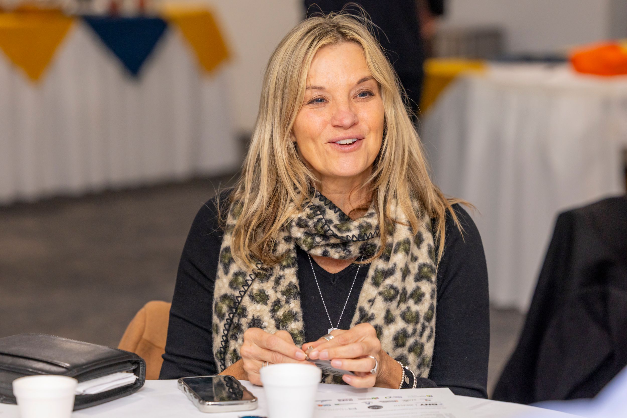 A person in a black top and leopard print scarf smiles while holding a coffee cup at an indoor event.
