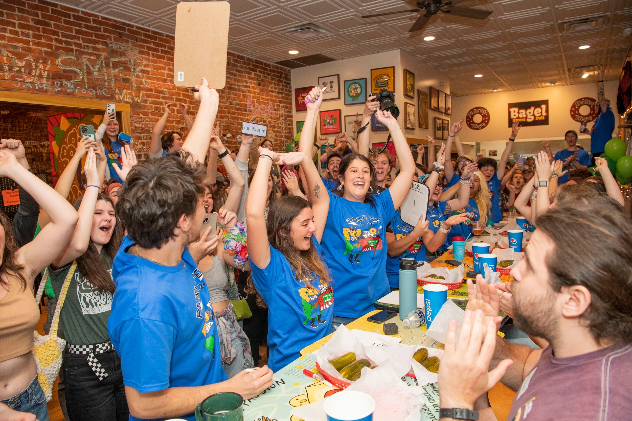 A large group of people in blue shirts cheer and raise their arms while seated at restaurant tables during a celebration.