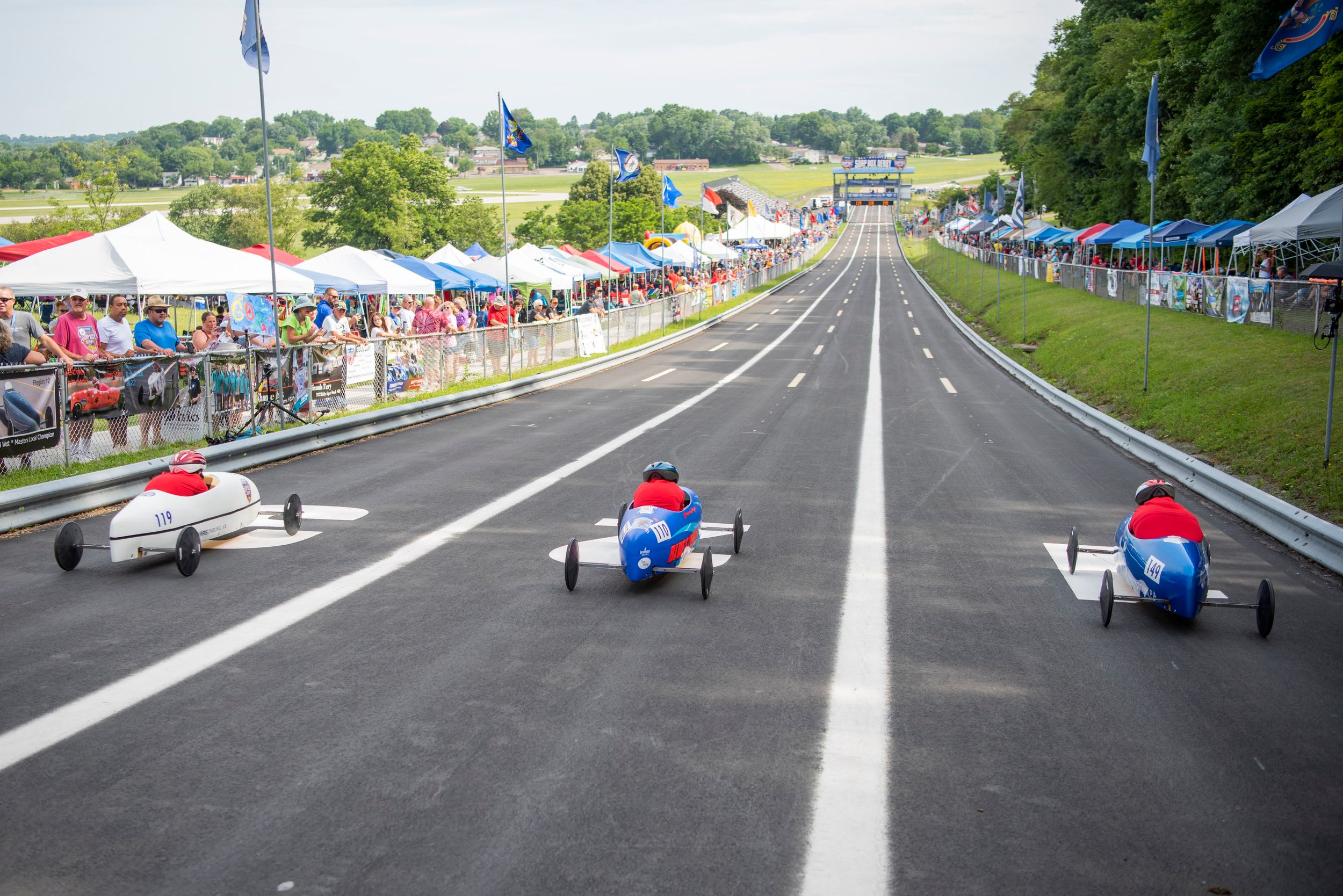 Vintage soapbox derby racers compete down a hill track with spectators lining both sides of the course.