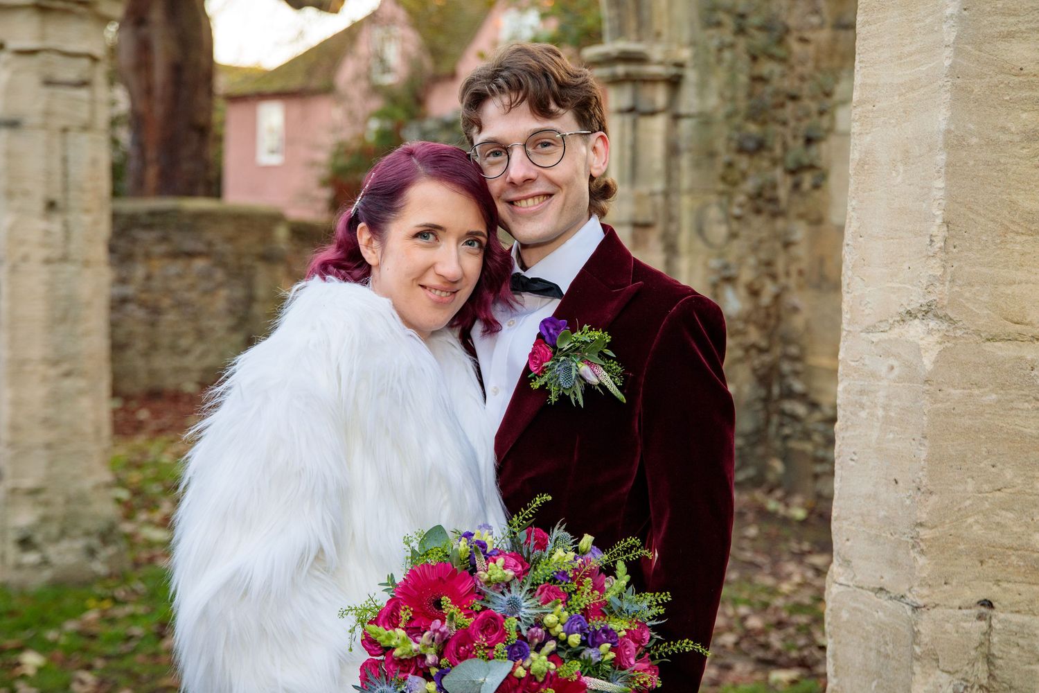 Bride and groom share romantic moments against stone wall backdrop with colorful floral bouquet.