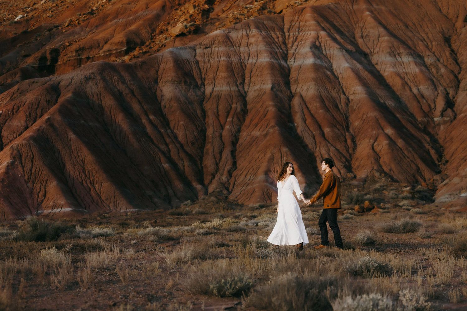 Couple holding hands walking through desert with striking red striped mountain backdrop.
