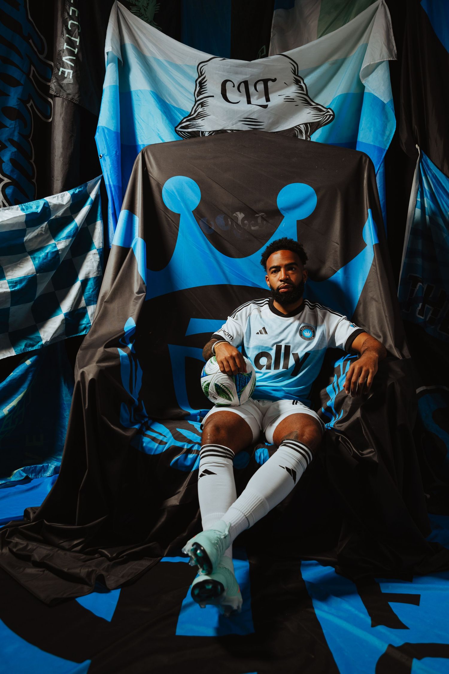 A Charlotte FC player sits confidently in a chair wearing a white Adidas ally jersey with blue flags.