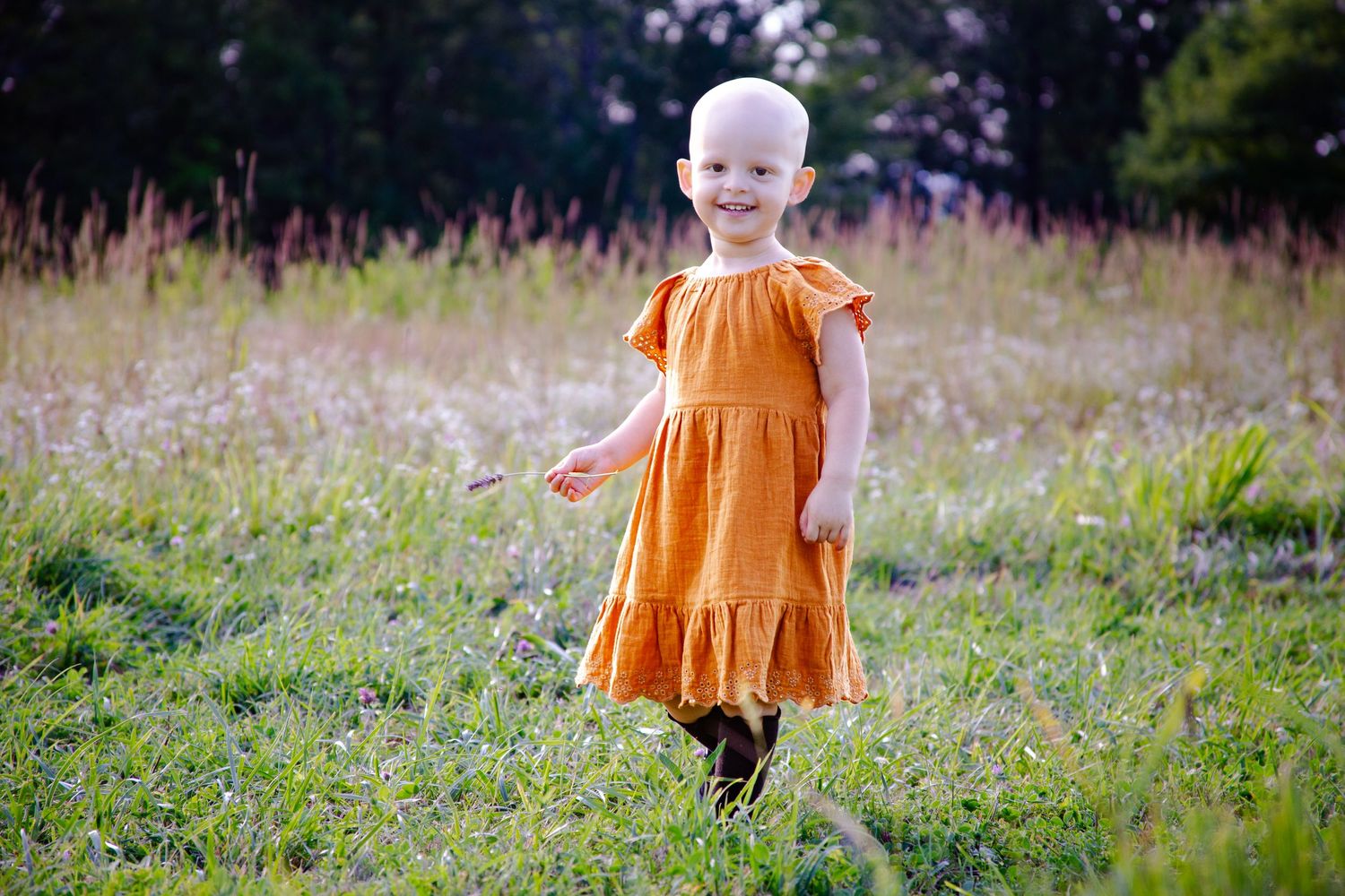 Small child in orange dress twirls playfully in a sunlit meadow filled with wildflowers.