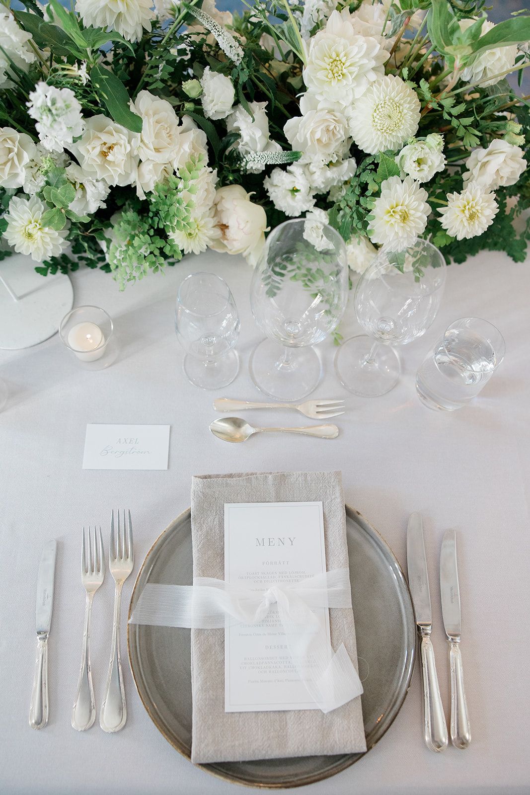 Elegant wedding table setting with white floral arrangements, crystal glassware, silver cutlery and gray charger plates.