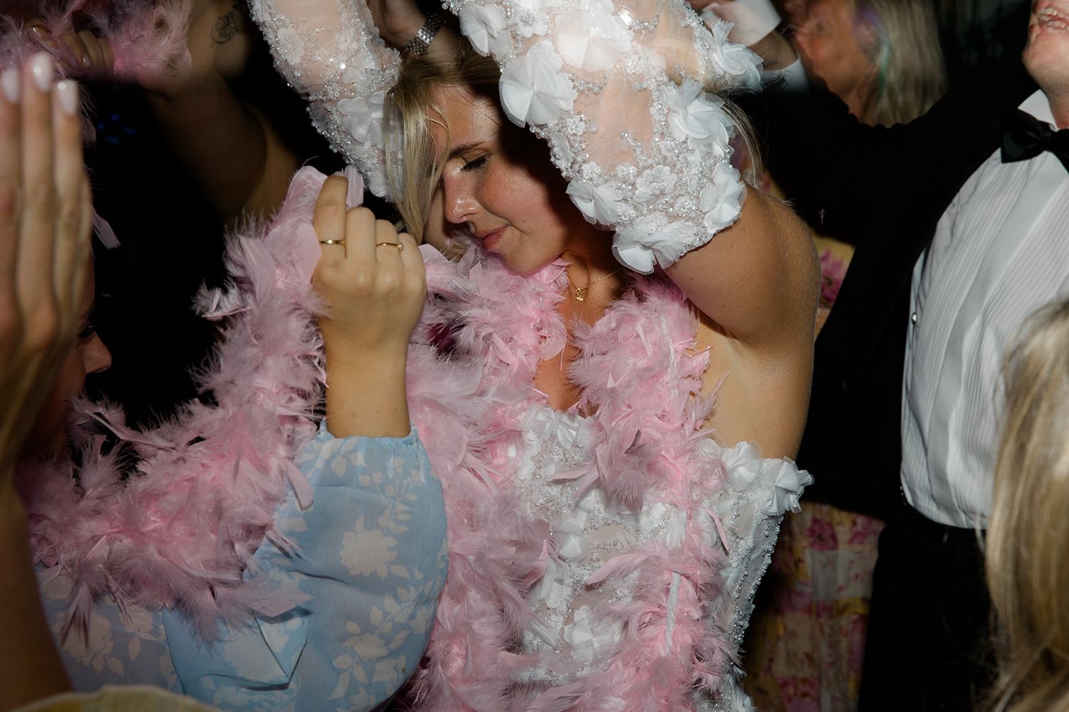 Party goers dance and celebrate wearing pink and white feather boas at a festive nighttime event.