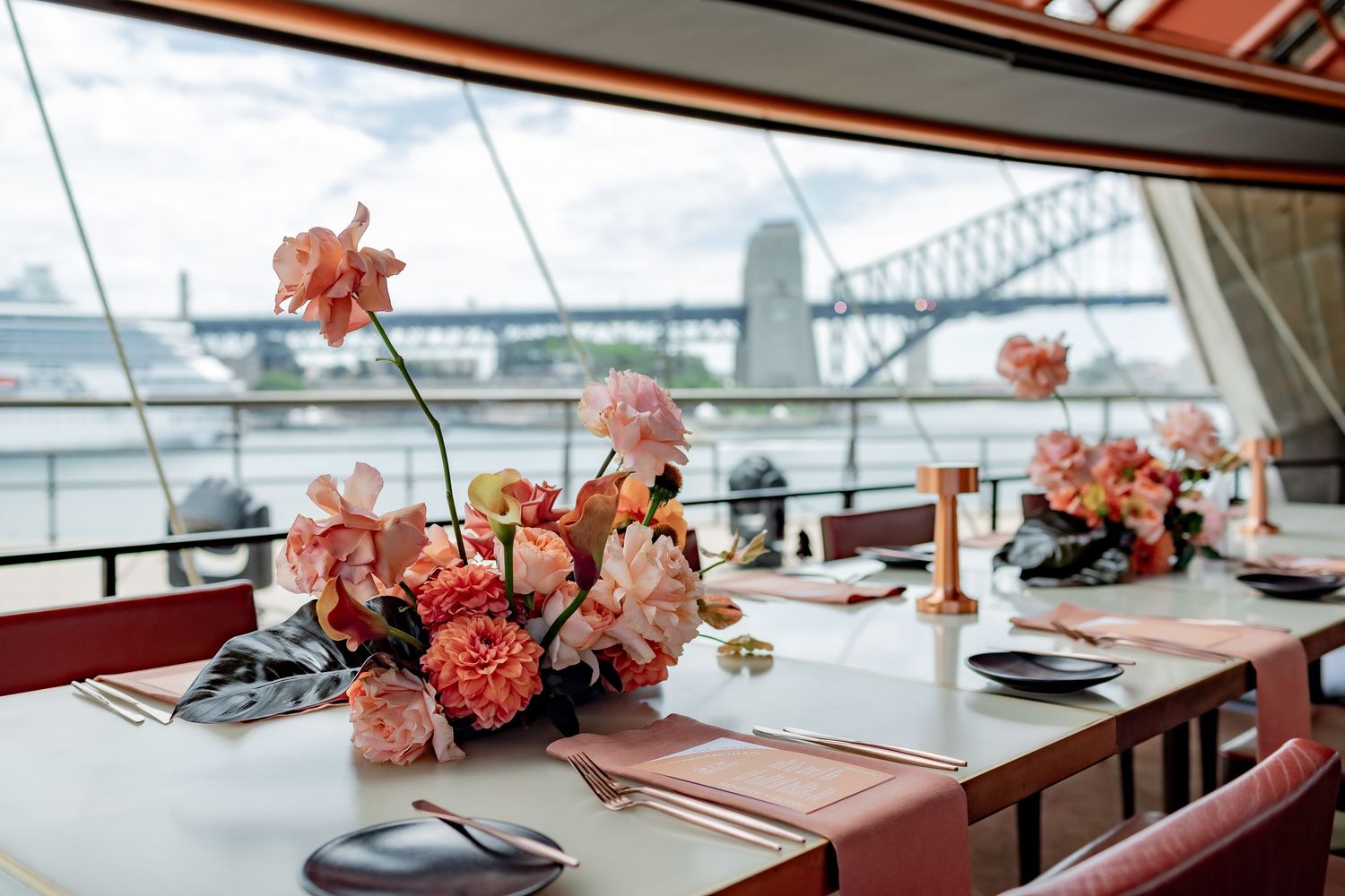 Elegant restaurant table settings with pink floral centerpieces overlooking a waterfront view through large windows.