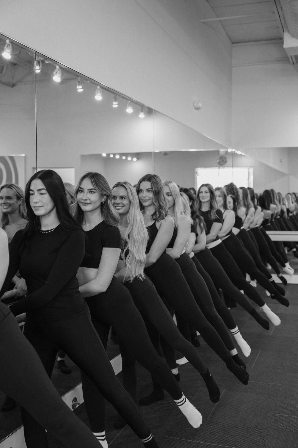 A line of dancers in black attire perform a synchronized dance routine in a bright studio space.