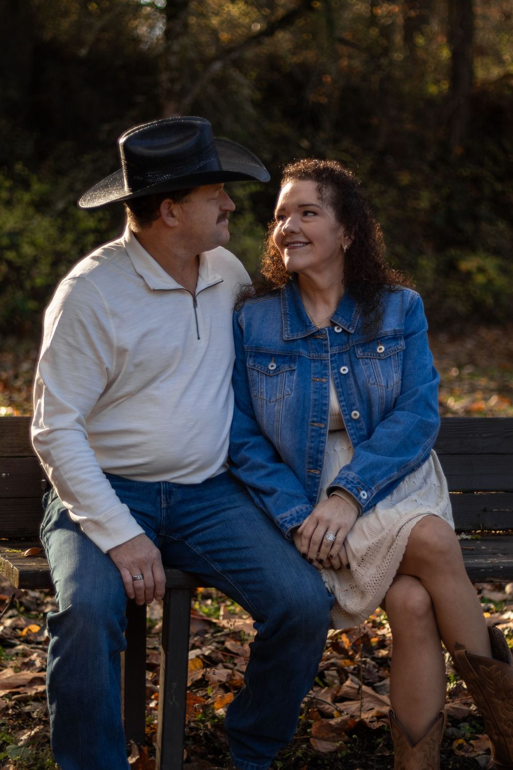 Couple sitting on bench in fall park, man in black cowboy hat and white shirt, woman in denim jacket smiling.