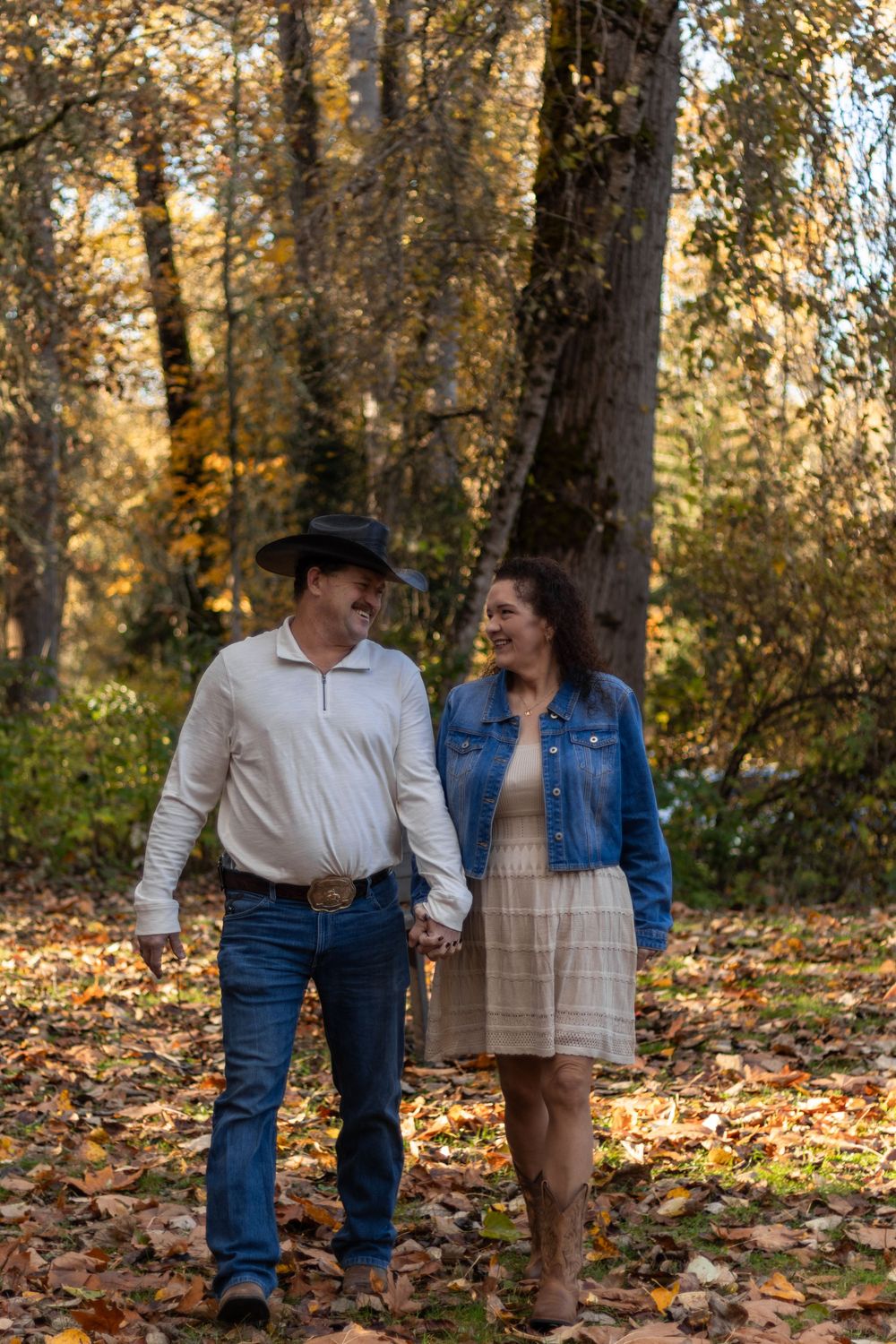 Couple walking hand in hand through autumn forest, man in black cowboy hat, woman in denim jacket and dress.