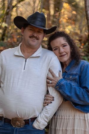 Couple posing in autumn forest, man in black cowboy hat and white zip pullover, woman in denim jacket embracing him.