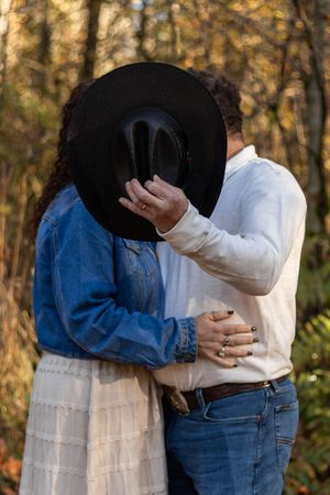 Couple sharing a hidden kiss behind black cowboy hat in golden autumn forest setting.